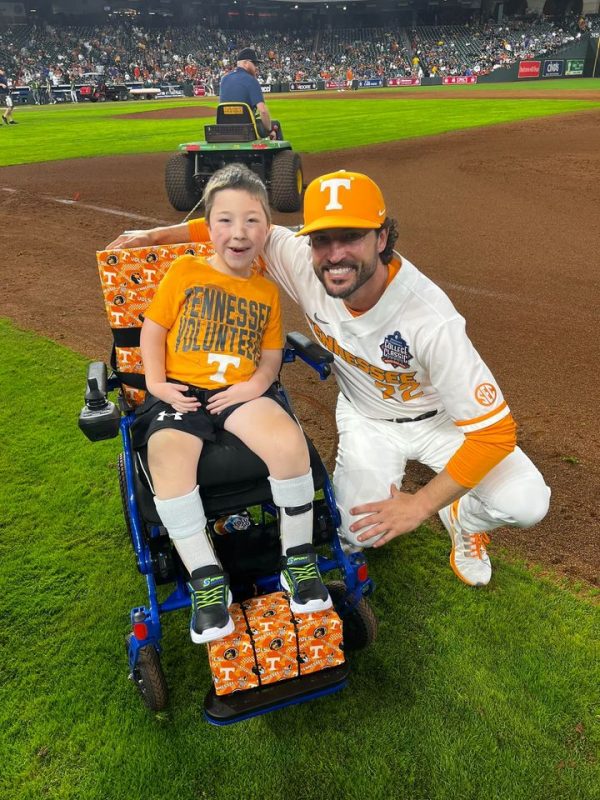 8 year old boy in a wheelchair with a big smile wearing an orange Vols t-shirt and a Tennessee Vols baseball player crouched  next to him with a big smile, too!